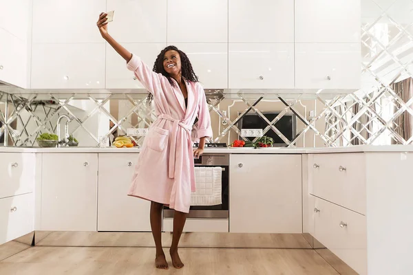 幸せな黒人女性が台所で selfie を作るします。女性が身に着けているピンクのバスローブ、暗くて長い巻き毛. — ストック写真