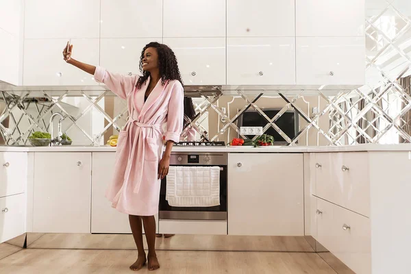 魅力的な幸せ黒人女性台所に立って、selfie を作るします。ピンクのバスローブ、長い巻き毛を身に着けている女性. — ストック写真