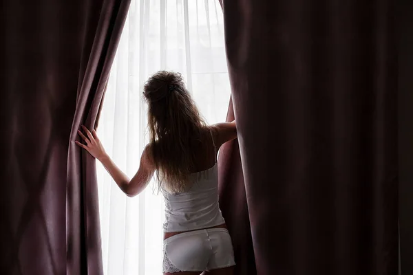 Mulher branca esbelta abrindo as cortinas e olhando pela janela. Mulher tem cabelo encaracolado longo e pijamas vestindo . — Fotografia de Stock