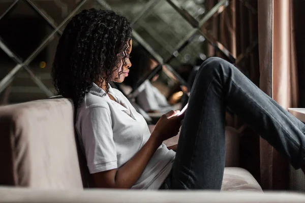 Rychlý vzkaz pro přítele. Atraktivní mladá Afričanka drží chytrý telefon a podíváme se na to při sezení na pohovce. — Stock fotografie