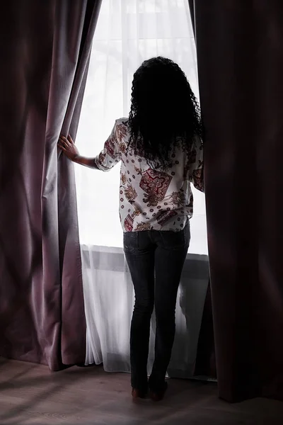 Belle femme noire avec de longs cheveux bouclés foncés debout près de la fenêtre et regardant par elle . — Photo