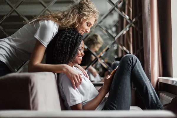 Two young attractive women - black and white, looking at the screen of the phone. Living room location. — Stock Photo, Image