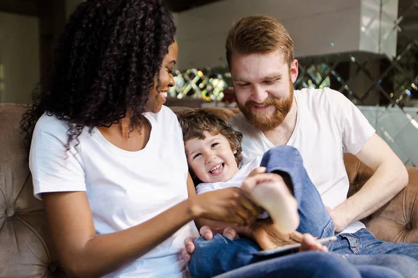 Happy family of black woman and white man sitting on the sofa with their son and laughing.