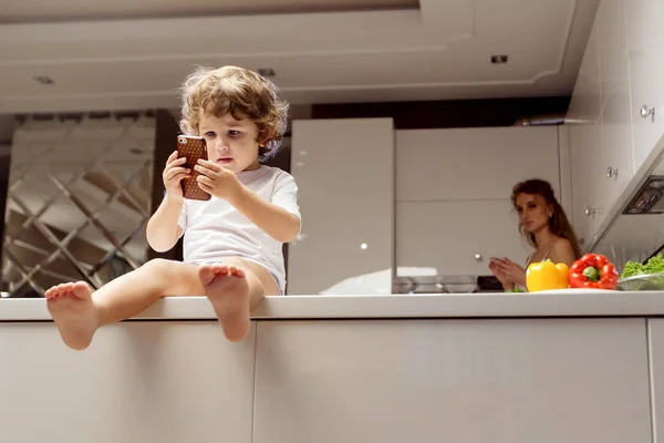 Menino sentado na superfície da cozinha com o telefone nas mãos . — Fotografia de Stock