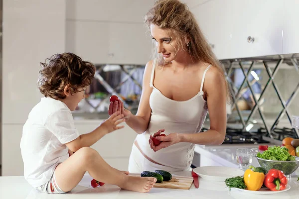 Mother showing tomatoes to her little son. Moder white kitchen location. — Stock Photo, Image