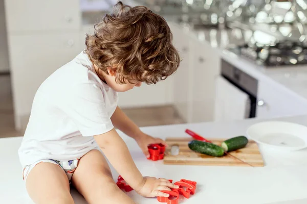 Little boy sitting on the kitchen sufface playing with vegetables. — Stock Photo, Image