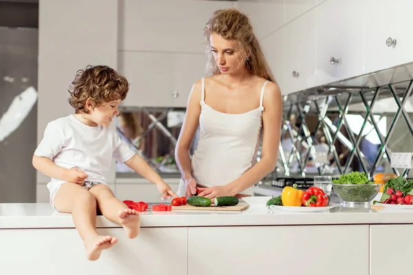 Attractive mother making a salad with her little son. Boy sitting on the kitchen surface. White modern kitchen location. — Stock Photo, Image