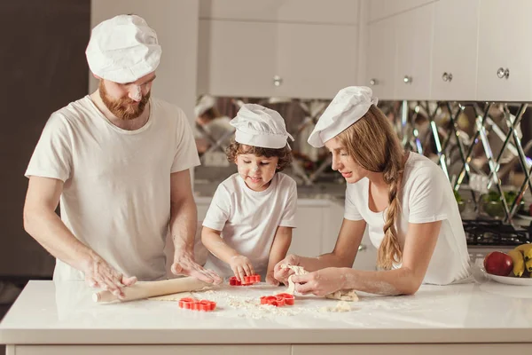 Happy family of father, mother and little son making dough together in the modern white kitchen. Family wearing white aprons and hats. — Stock Photo, Image