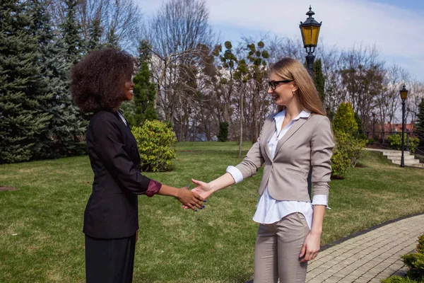 Twee jonge aantrekkelijke businesswomenn schudden handen na goede deal. Een vrouw is zwart. Vrouwen permanent buiten. — Stockfoto