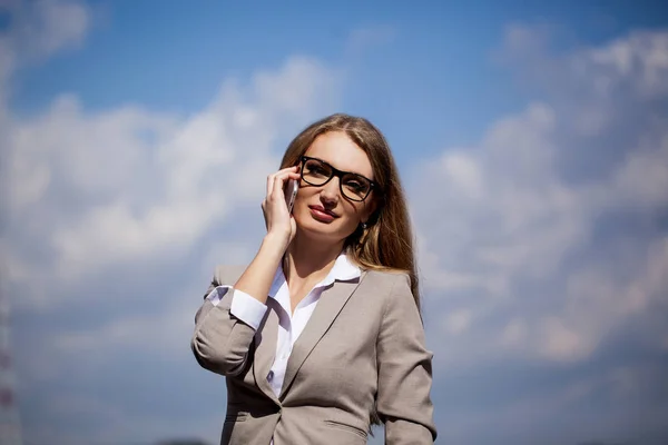 美丽的女企业家站在天空背景，谈电话的肖像。女人穿着西装和眼镜. — 图库照片