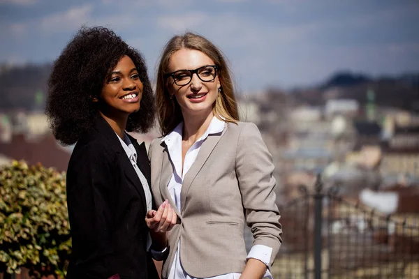 Mulheres de negócios sorridentes em ternos posando para câmera no parque com ótima vista para a cidade. Mulher negra tem cabelo encaracolado . — Fotografia de Stock