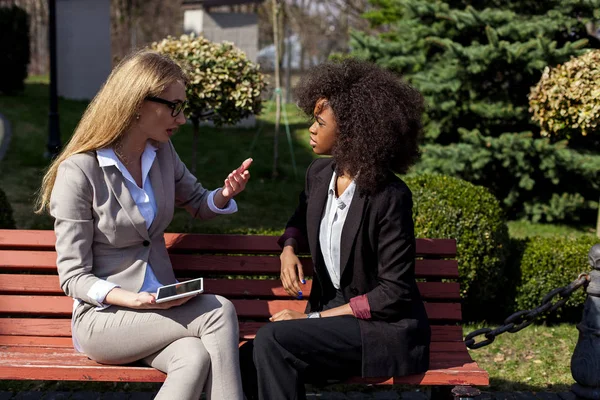 Twee aantrekkelijke jonge vrouwen in de kostuums zitten in het park en het gesprek. Een vrouw is afroamerican, — Stockfoto