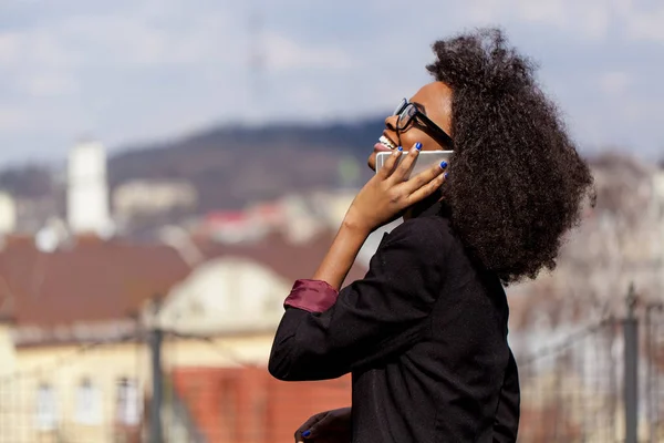 Черная деловая женщина разговаривает по телефону и улыбается. Отличный вид на город . — стоковое фото