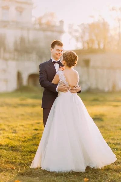 The emotional outdoor photo of the hugging newlyweds during the sunset. The back view of the bride with the bare shoulders. — Stock Photo, Image