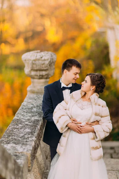 На осенней фотографии жених обнимает невесту, стоя на готической лестнице на фоне пожелтевших деревьев. . — стоковое фото