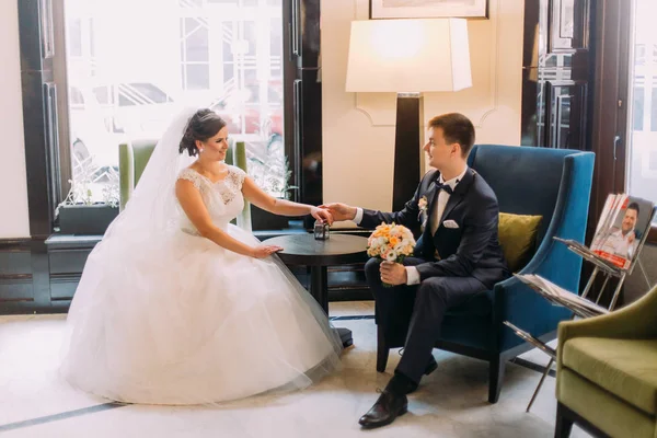 Retrato de noivo jovem elegante recém-casado vestindo terno azul e noiva encantadora em vestido lindo. O par sentado em cadeiras com fundo luxuoso — Fotografia de Stock