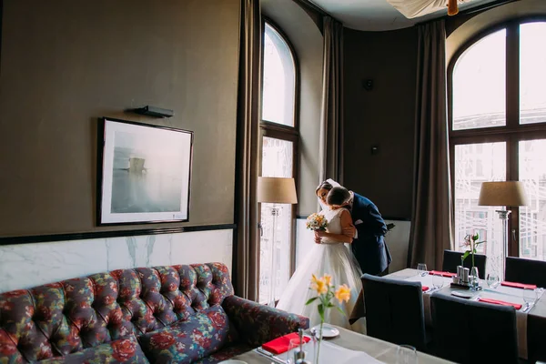 Beautiful, romantic newlyweds near bright window in wedding hall before the celebration. Groom is kissing his bride — Stock Photo, Image