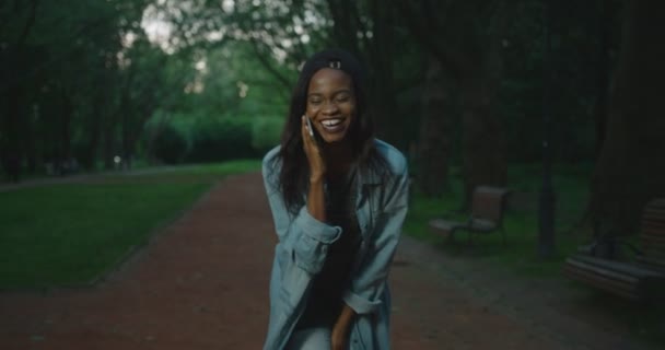 Sorrindo afro-americana menina está rindo enquanto conversa via telefone durante a caminhada no parque da noite . — Vídeo de Stock