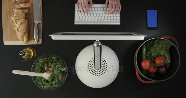 O homem está sentado em frente ao computador na cozinha. Legumes e pão estão sobre a mesa. Vista superior . — Vídeo de Stock