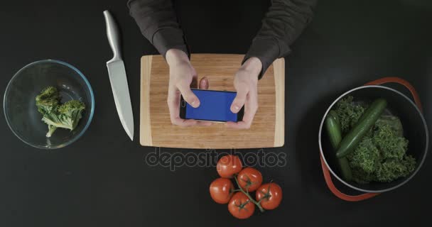 Κάτοψη του χέρια κρατώντας το τηλέφωνο πάνω από τον πίνακα. Έννοια κουζινών. Ξύλο κοπής, μαχαίρι και λαχανικά που τοποθετείται στον πίνακα. — Αρχείο Βίντεο