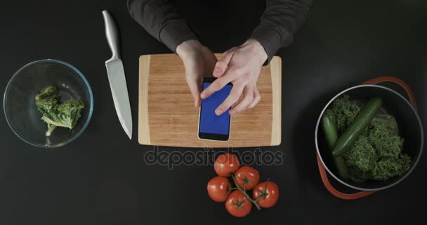 Vista superior de las manos desplazándose por la pantalla del teléfono. Manos sosteniendo el teléfono por encima de la tabla de cortar y comida . — Vídeos de Stock