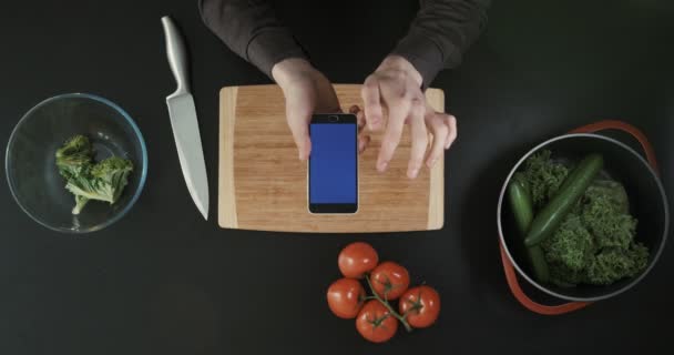 Вид зверху. Руки, що тримають телефон з синім екраном. Овочі, обробна дошка і ніж на столі . — стокове відео