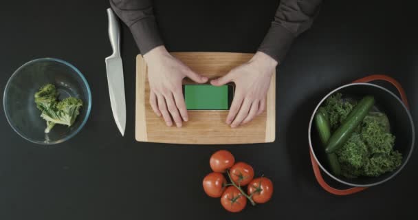 Ręce, trzymając telefon nad drewnianą deską do krojenia w pobliżu żywności i wielki nóż. Widok z góry. Zielony ekran. — Wideo stockowe