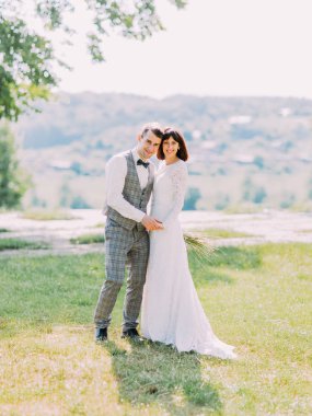Gülümseyen yeni evliler el ele tutuşarak ve çayır üzerinde kafa kafaya ayakta çok güzel tam uzunlukta portresi.