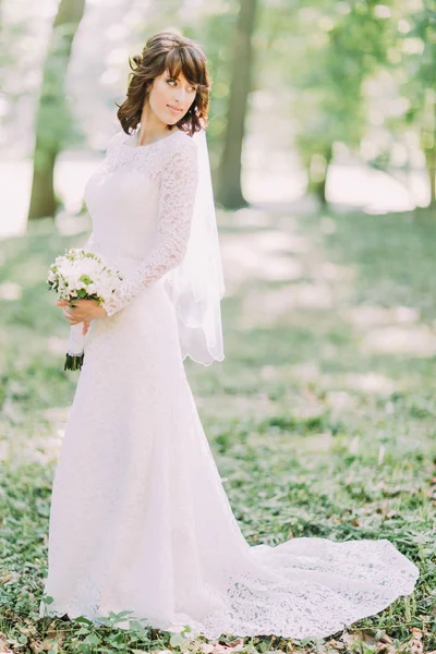 La novia con el ramo de bodas está mirando al lado izquierdo en el fondo del parque . — Foto de Stock