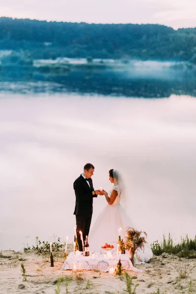 Fullängds beskåda av de härliga nygifta hålla skinande ljuset under en romantisk picknick på bakgrunden till floden och skogen. — Stockfoto