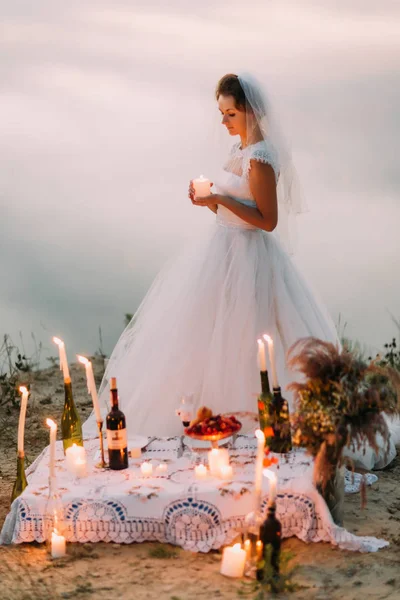A foto completa da noiva elegante segurando a vela brilhante atrás do conjunto de piquenique romântico . — Fotografia de Stock