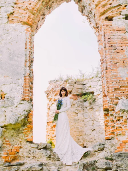 Fullängds beskåda av tankeväckande bruden med blommorna står på ruinerna av huset. — Stockfoto