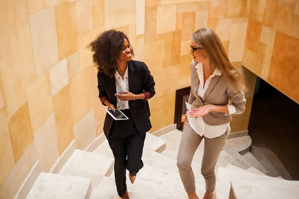 Shot of two smiled businesswomen going upstairs. Women wearing suits. One woman is black. — Stock Photo, Image