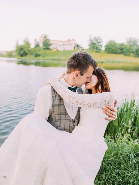 Das Nahaufnahme-Außenporträt des Bräutigams, der die Braut im Hintergrund des Flusses trägt und küsst. — Stockfoto