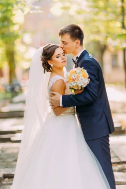 Damat gelini güneşli parkta kafasına öpüyor. Hassas atmosfer.