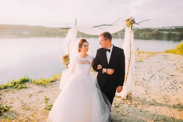 La alegre pareja de recién casados está caminando brazo en brazo en el fondo del arco decorado y el río . — Foto de Stock