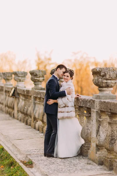 O noivo está abraçando a noiva no vestido longo inclinando-se sobre a cerca antiga do palácio . — Fotografia de Stock