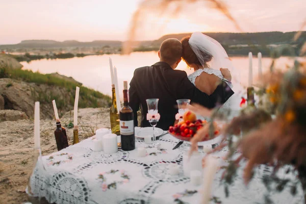 La coppia di sposi appena sposati si sta godendo il tramonto vicino al fiume dietro il set da picnic . — Foto Stock