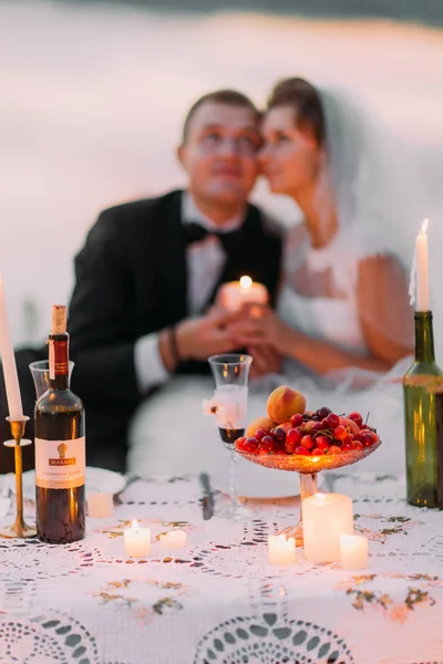 La vista ravvicinata del set da picnic decorato con frutta, vino e candele brillanti sullo sfondo sfocato della felice coppia di sposi . — Foto Stock