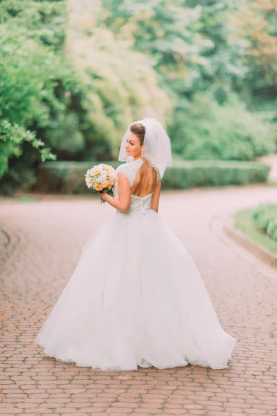 Полный вид на невесту с обнаженным плечом, держащую свадебный букет на заднем плане парка . — стоковое фото