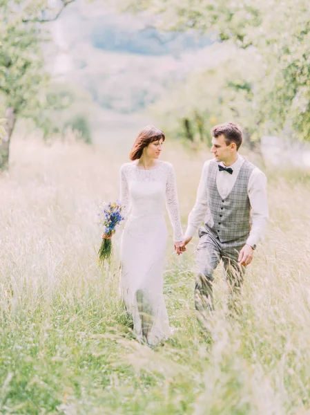 La belle promenade des jeunes mariés heureux dans le domaine. La mariée tient le bouquet des fleurs du champ bleu . — Photo