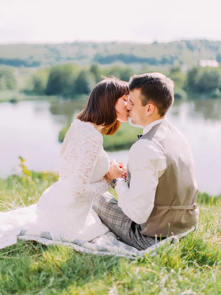 Счастливые молодожены держатся за руки и целуются, сидя на траве . — стоковое фото