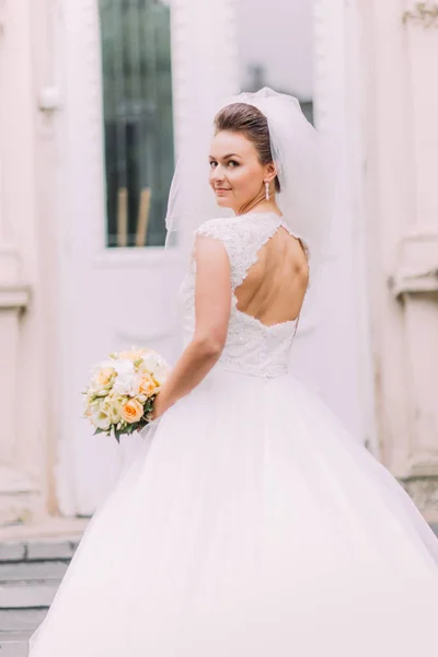 A vista de trás da noiva com as costas nuas. A noiva está olhando para a câmera enquanto segura o buquê de casamento . — Fotografia de Stock