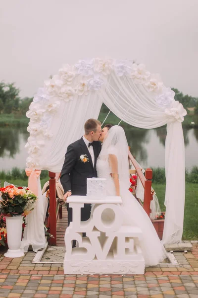 De full-length foto van de kussen pasgetrouwden achter het teken van de bruiloft en de boog. — Stockfoto