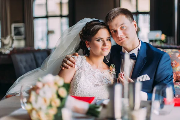 Enloved szczęśliwy newlywed para trzymając się nawzajem siedząc na kanapie. Luksusowe wnętrze jasne z dużymi oknami jako tło — Zdjęcie stockowe