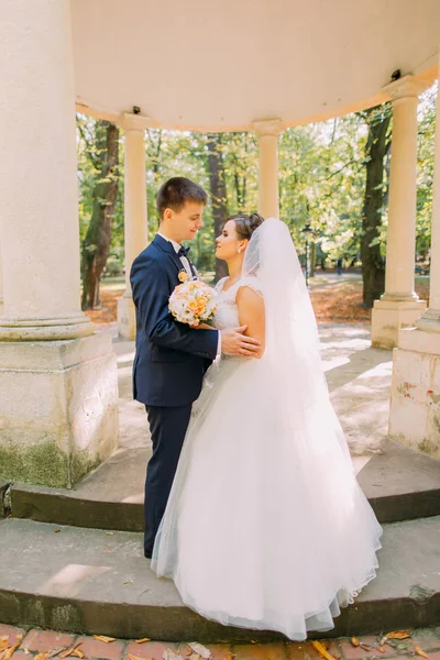 The hugging newlyweds standing near the old alcove at the background of the park. — Stock Photo, Image