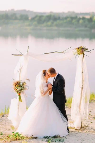 Celovečerní vertikální pohled líbání novomanželé během svatebního obřadu v blízkosti řeky. — Stock fotografie