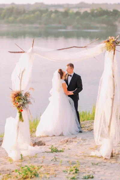 Los recién casados abrazos detrás del arco decorado cerca del río . — Foto de Stock