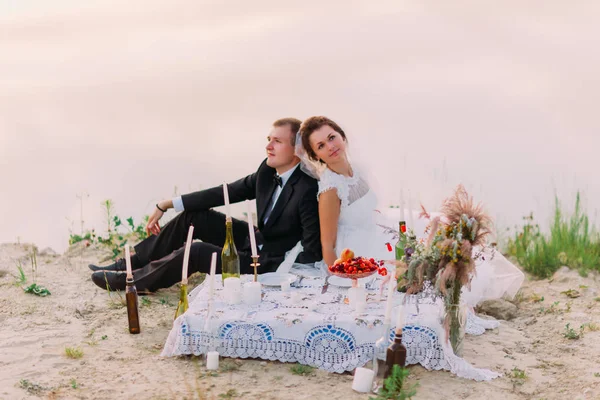 O retrato horizontal romântico dos recém-casados sirring back-to-back, tendo o piquenique de casamento na costa o piquenique de casamento na costa . — Fotografia de Stock
