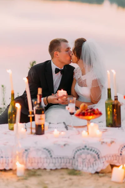 O casal recém-casado sentado está se beijando atrás do conjunto de piquenique decorado com velas brilhantes em garrafas . — Fotografia de Stock